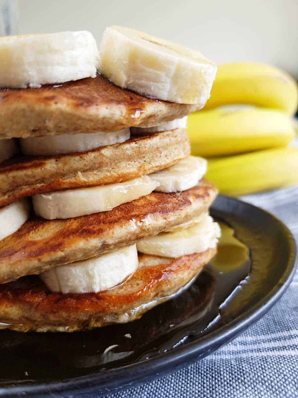 Stacked pancakes with banana slices and fresh bananas in the background.