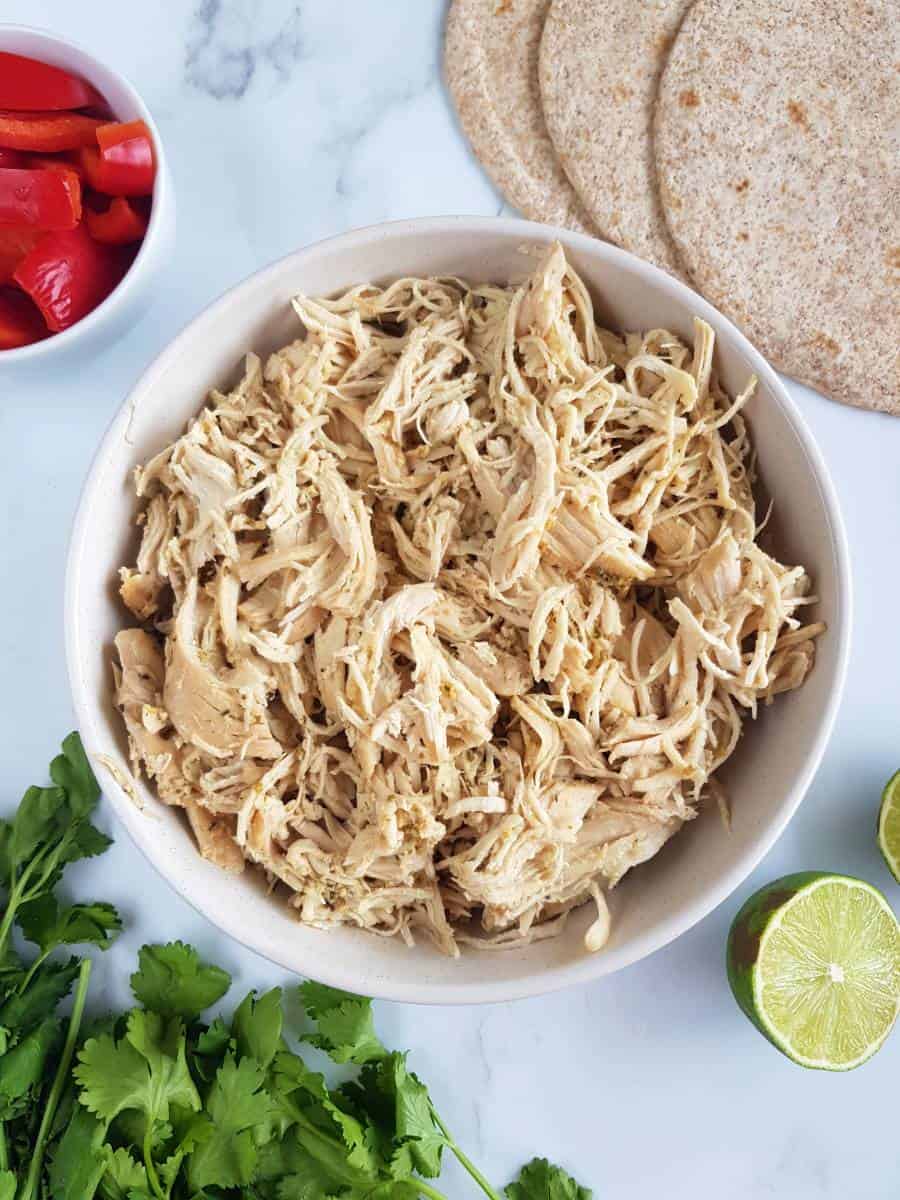 Crockpot mojo chicken in a bowl with cilantro, peppers and wraps on the side.