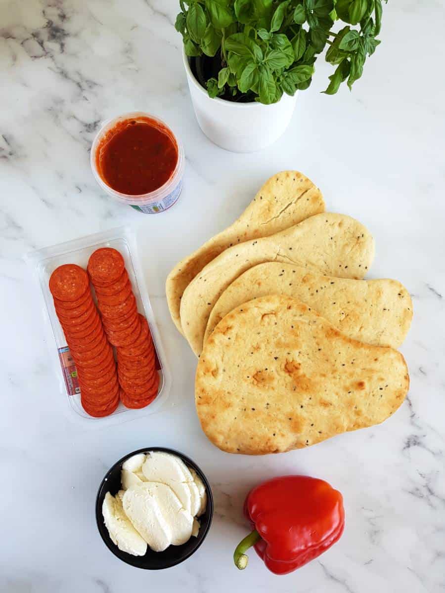 Naan pizza assembly table with naan breads, pepperoni, mozzarella, pizza sauce and basil.