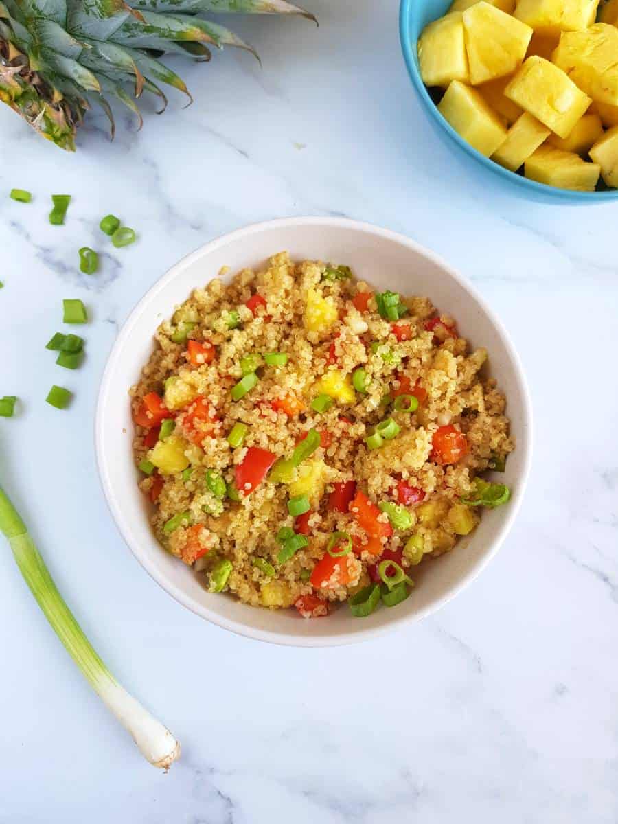 Pineapple fried quinoa in a white bowl with pineapple and scallions on the side.