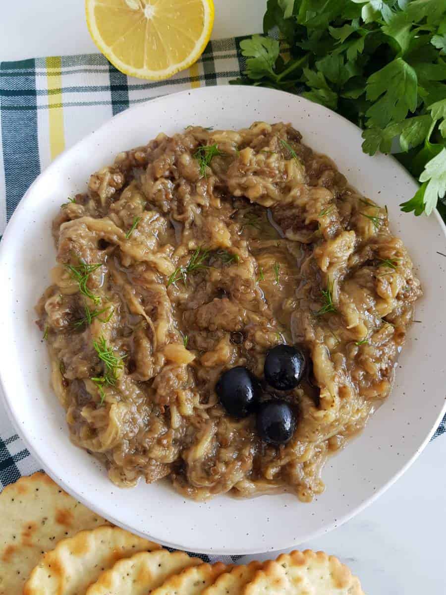 Greek eggplant dip in a bowl with parsley, lemon and crackers on the side.