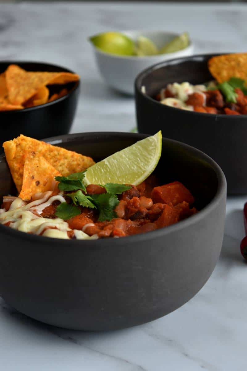 Mixed bean chilli in bowls, topped with lime wedge, tortilla chips and cheese.