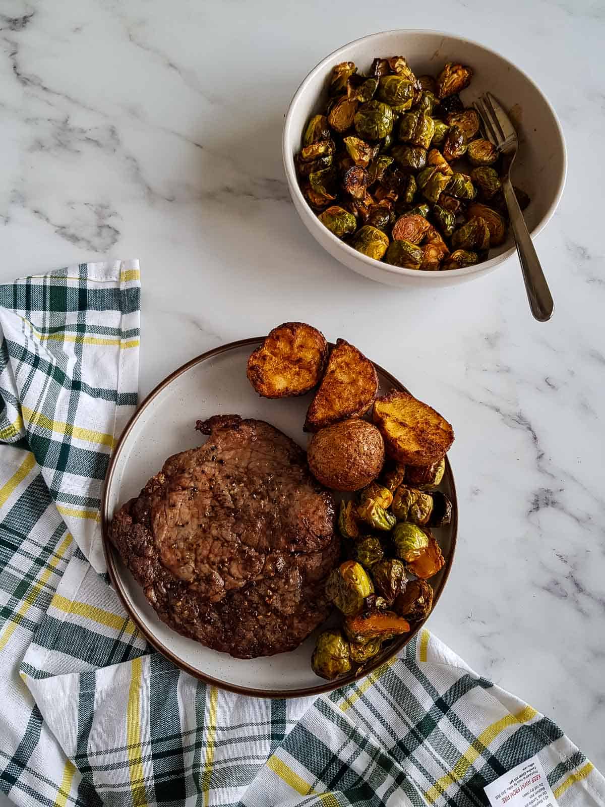 A plate of steak and side dishes, with a bowl of brussels sprouts in the back.