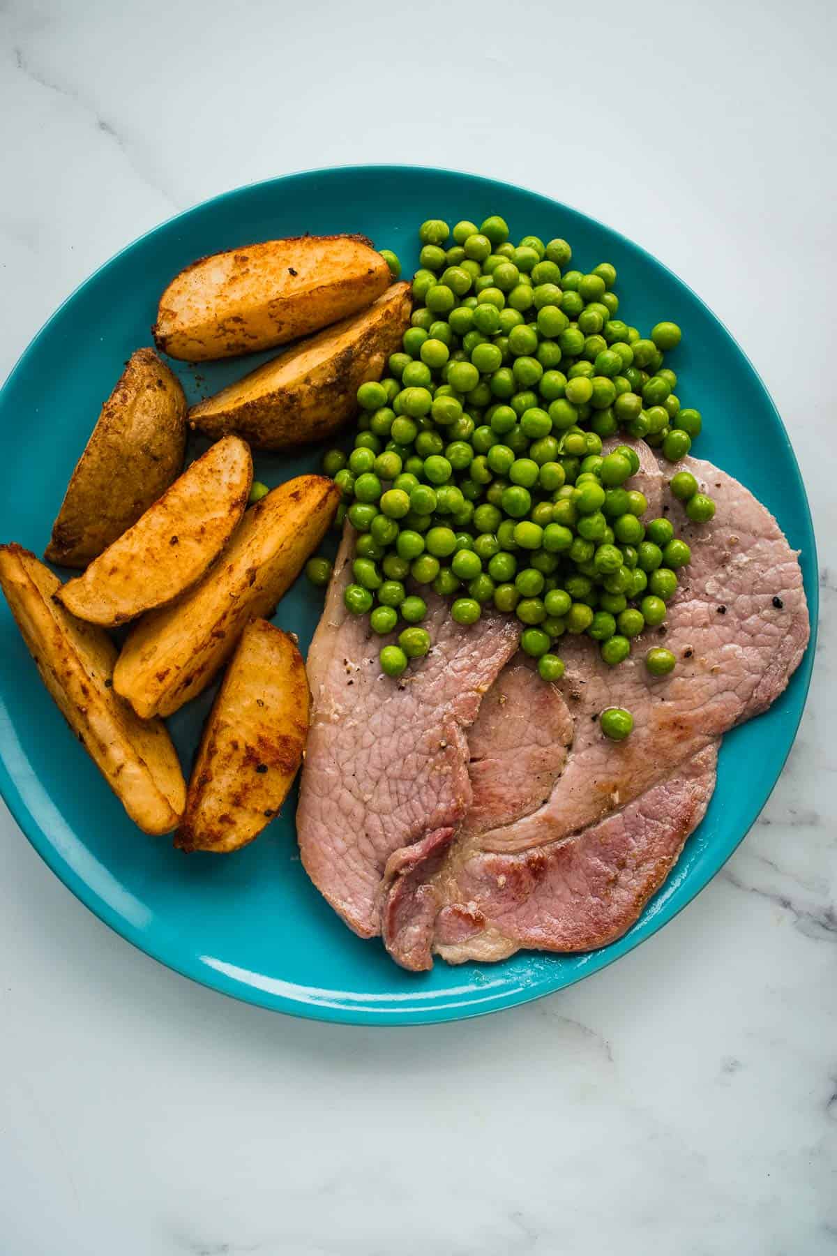 Gammon steaks on a plate with potato wedges and peas.