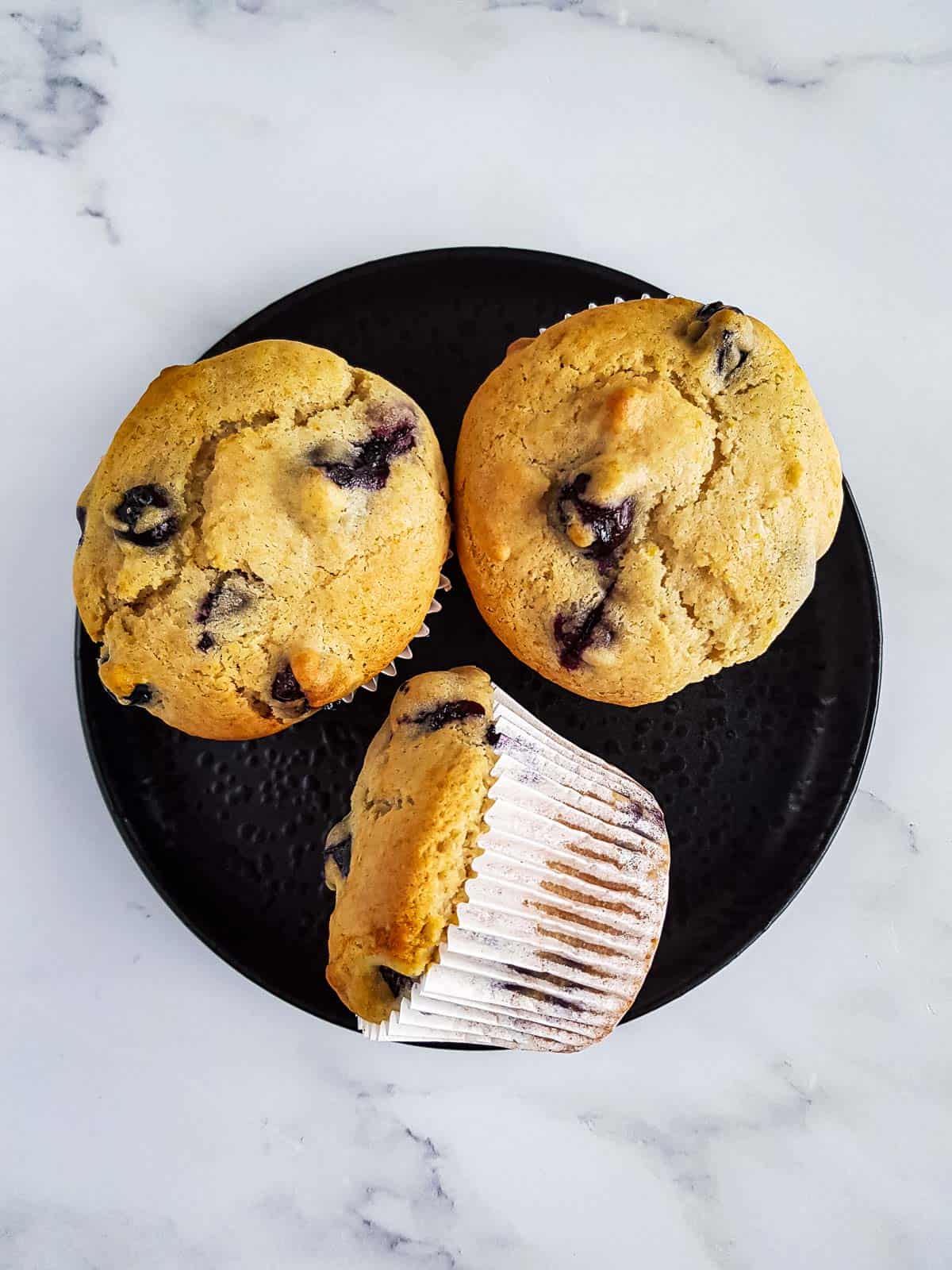 Three blueberry muffins on a plate, one is laid on the side.