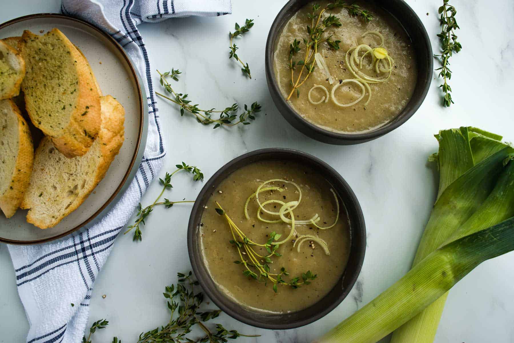 Bowls of soup in a table with a leek, garlic bread and fresh thyme.