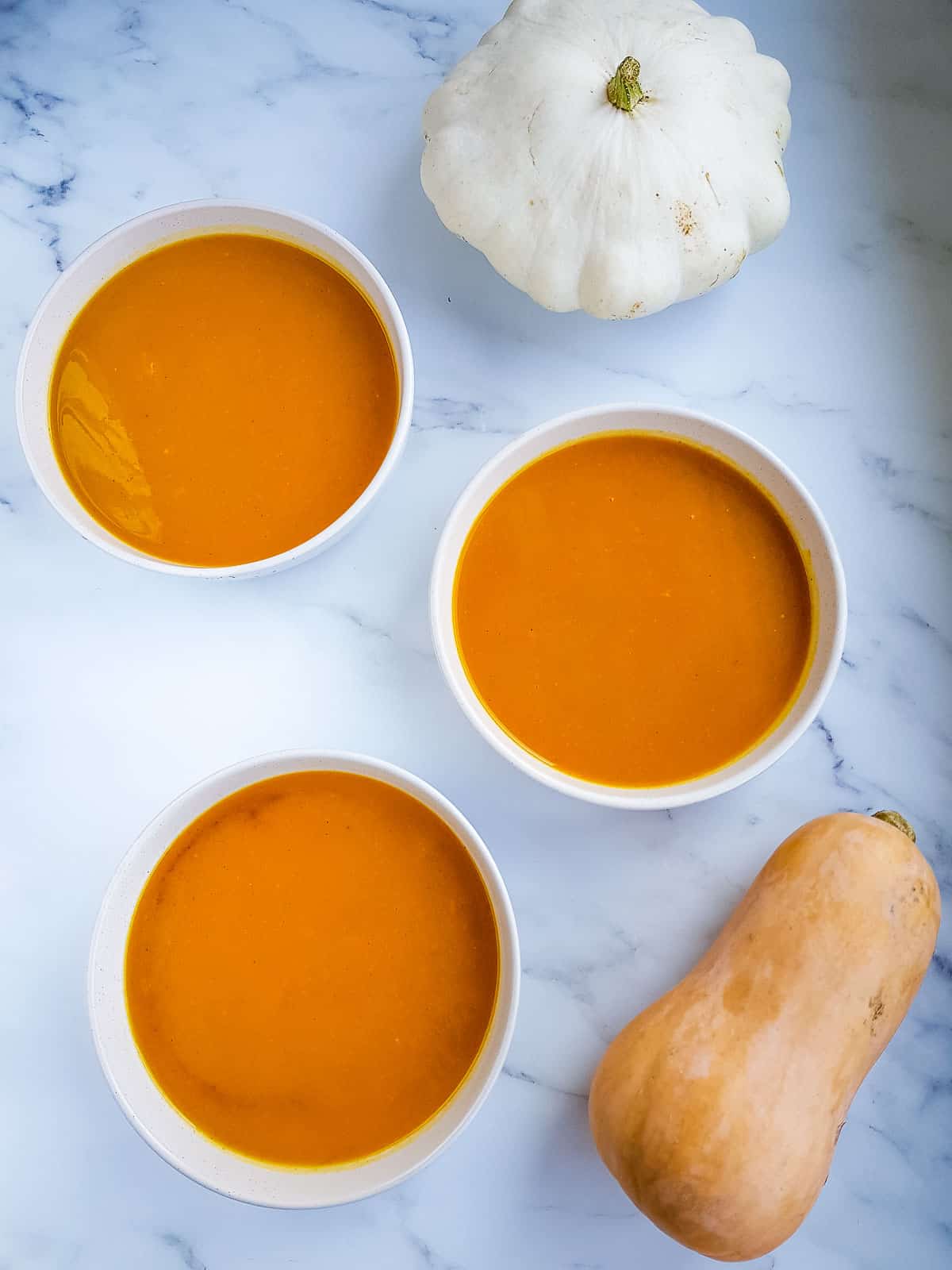 Three bowls of pumpkin soup with pumpkins on the side.