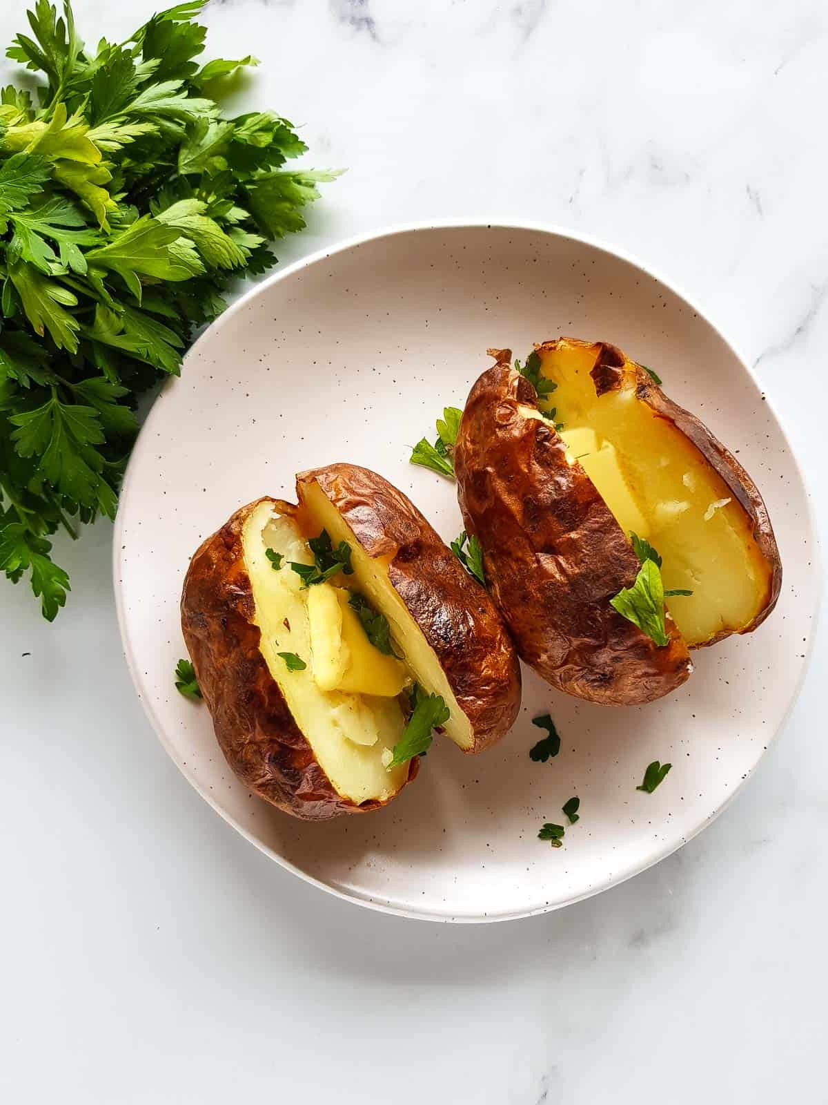 Two baked potatoes on a plate, with a bunch of parsley on the side.