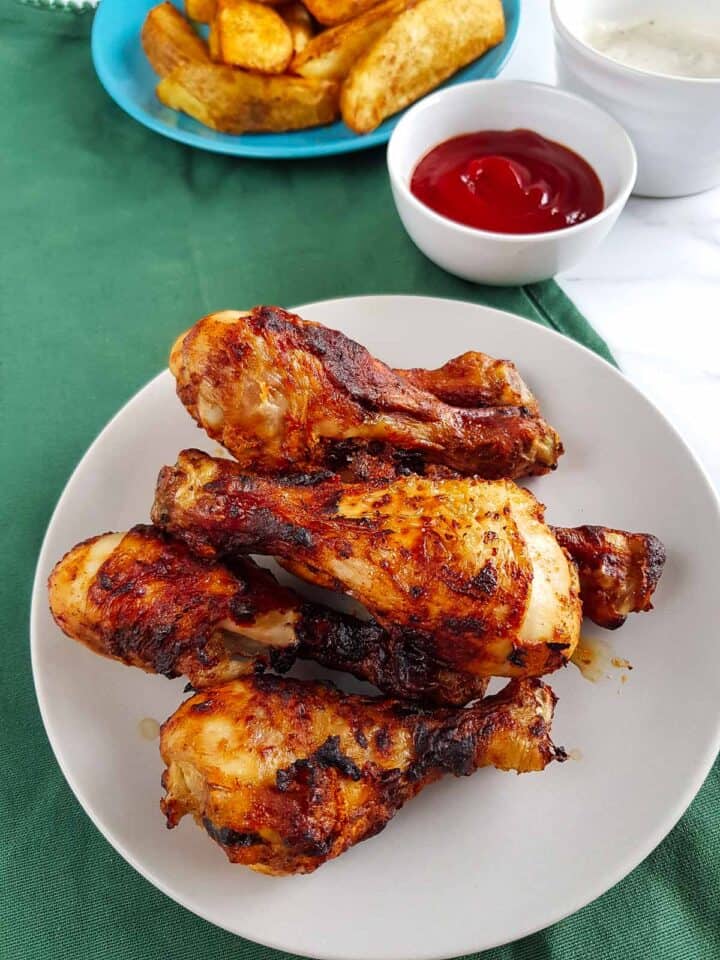 Air fried chicken drumsticks on a plate with potato wedges and ketchup in the background.