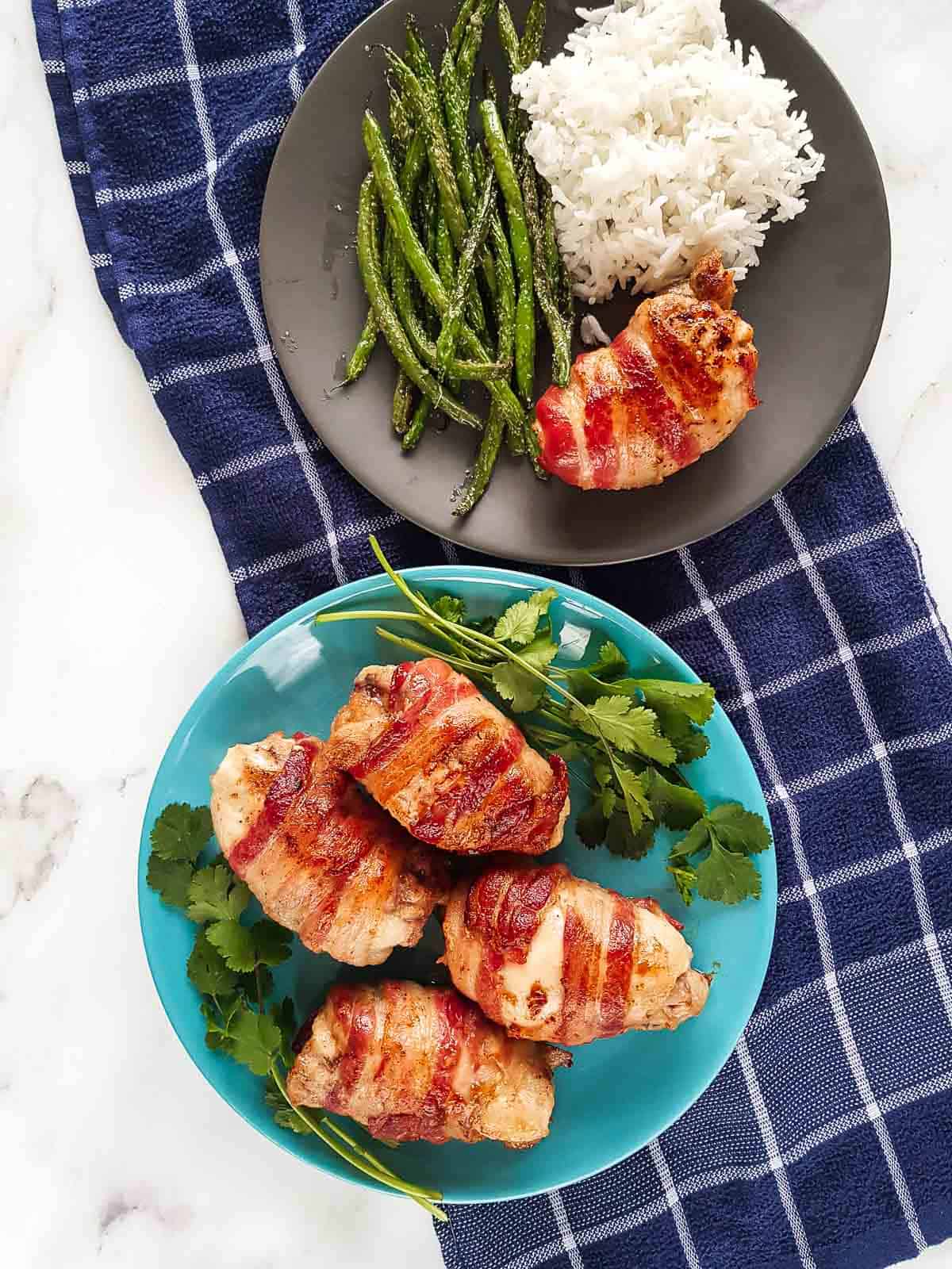 Bacon wrapped chicken on a plate with side dishes. Another plate with multiple chicken thighs on the side.