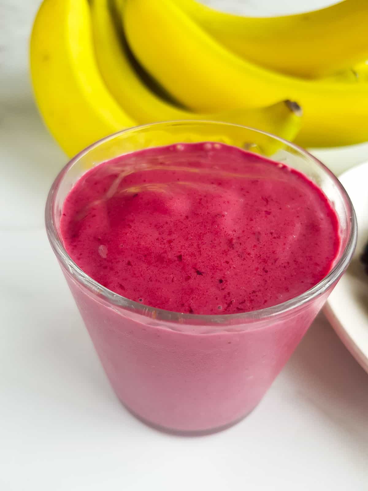 A glass of pink smoothie with bananas in the background.