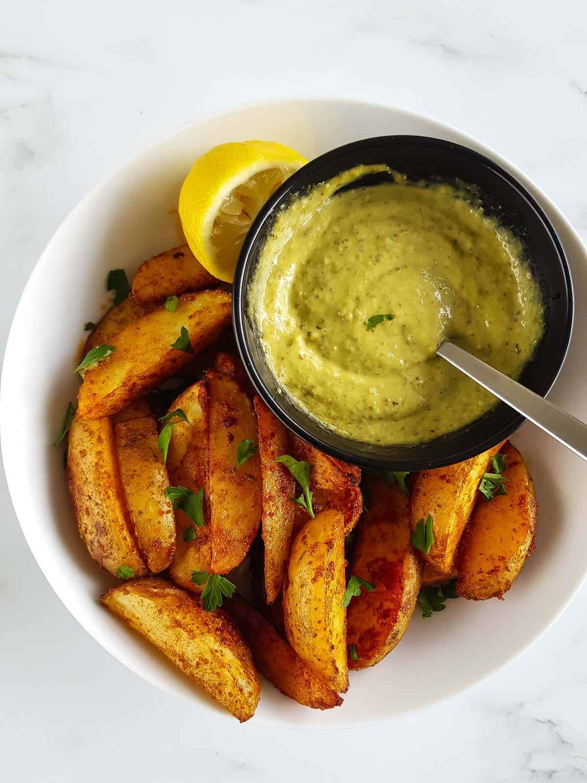 A plate of potato wedges, aioli with pesto and a lemon.