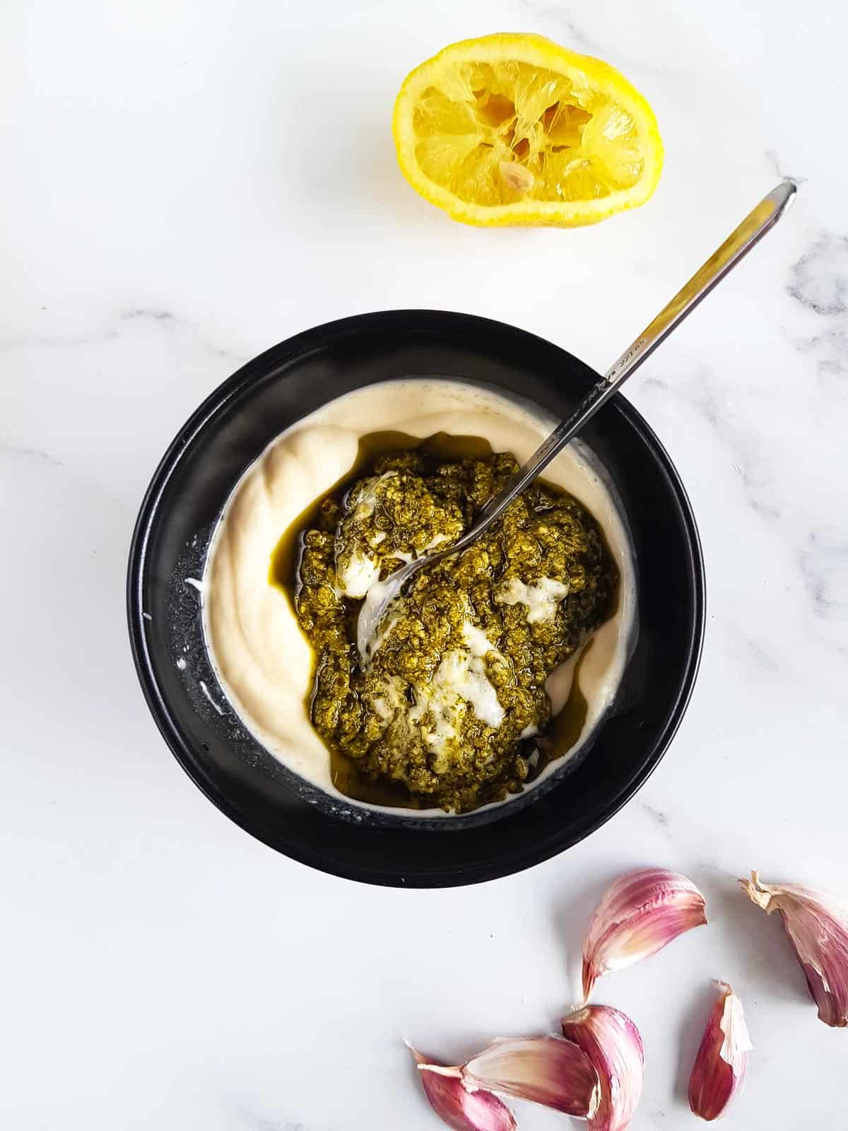 Mixing pesto and mayonnaise in a bowl, with garlic cloves and half a lemon on the side.