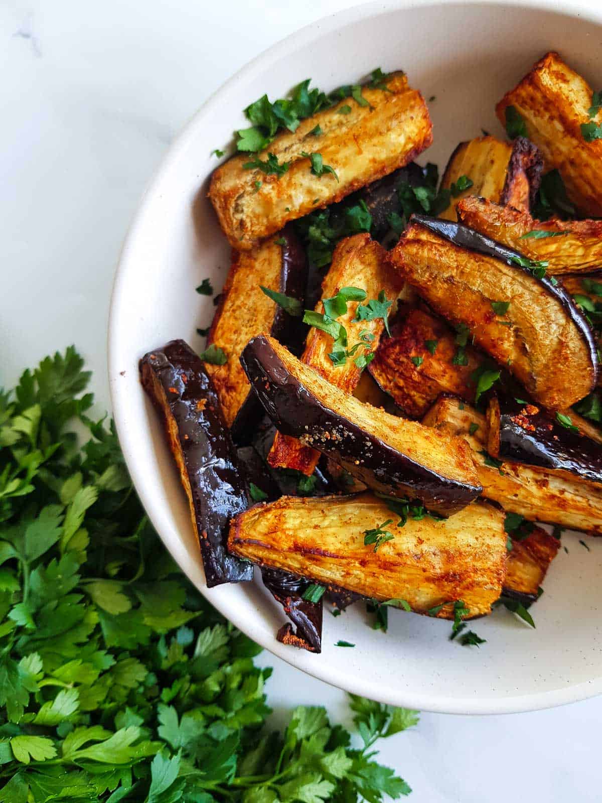 A white bowl of air fryer eggplant with parsley on the side.