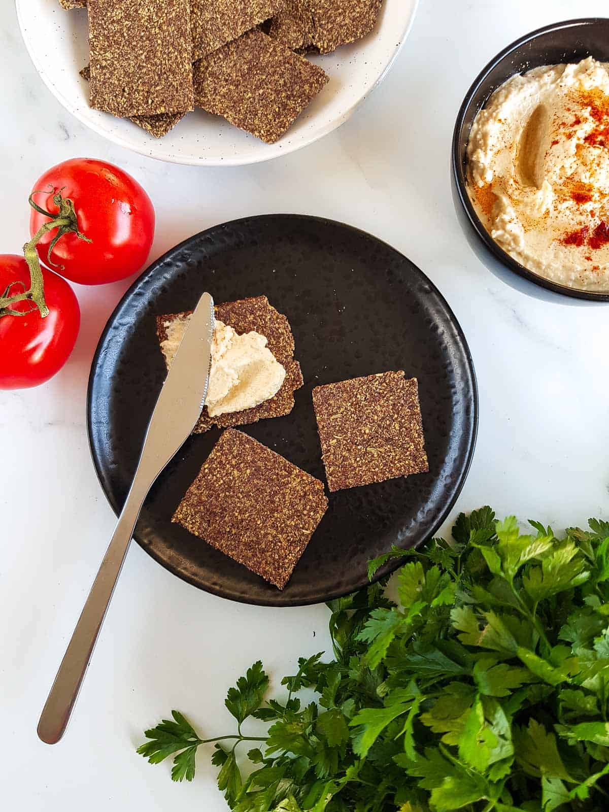 Crackers on a table with hummus, parsley and tomatoes on the side.