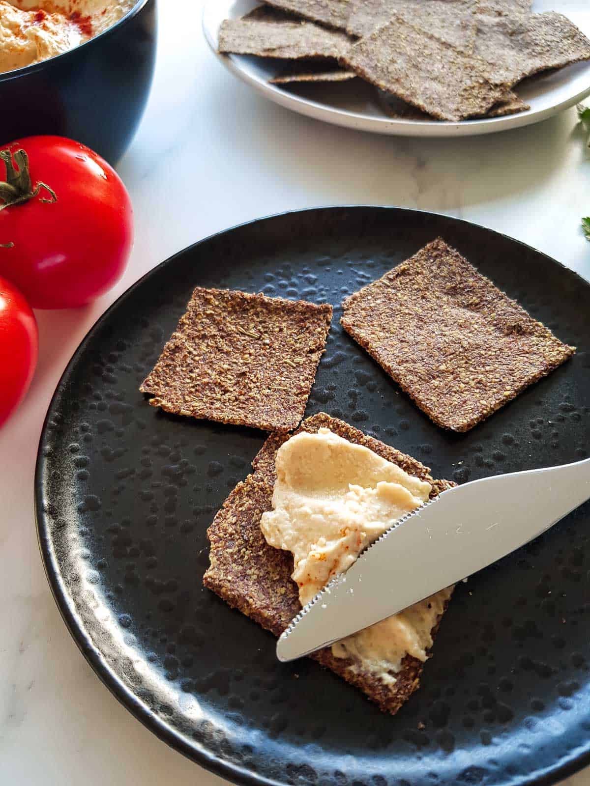 A knife spreading hummus onto a flax seed cracker.