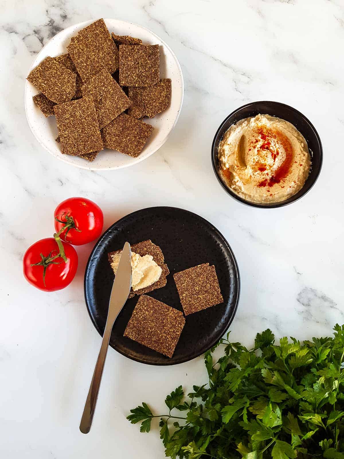 Flaxseed crackers on a plate, with a knife spreading hummus on top. A bowl of hummus and tomatoes on the side.