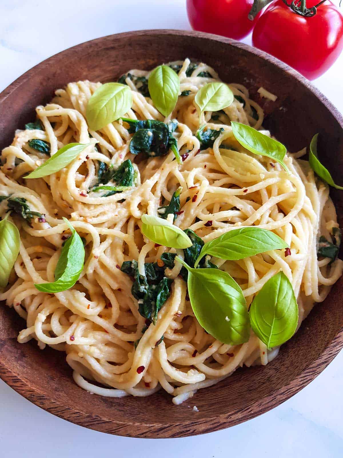 Pasta with hummus and spinach in a bowl, with tomatoes in the background.