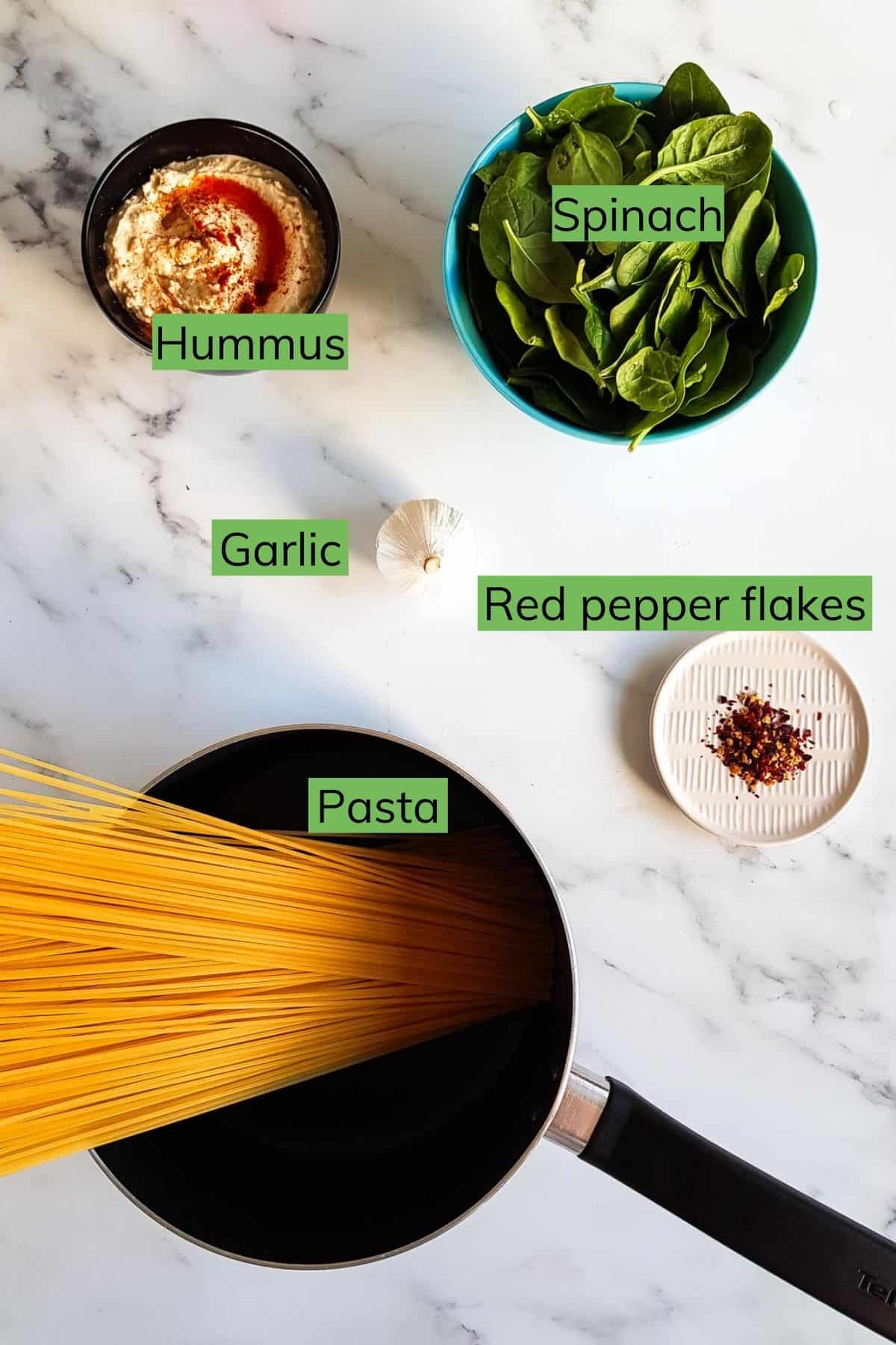 Garlic, spinach, hummus, red pepper flakes and pasta laid out on a table.