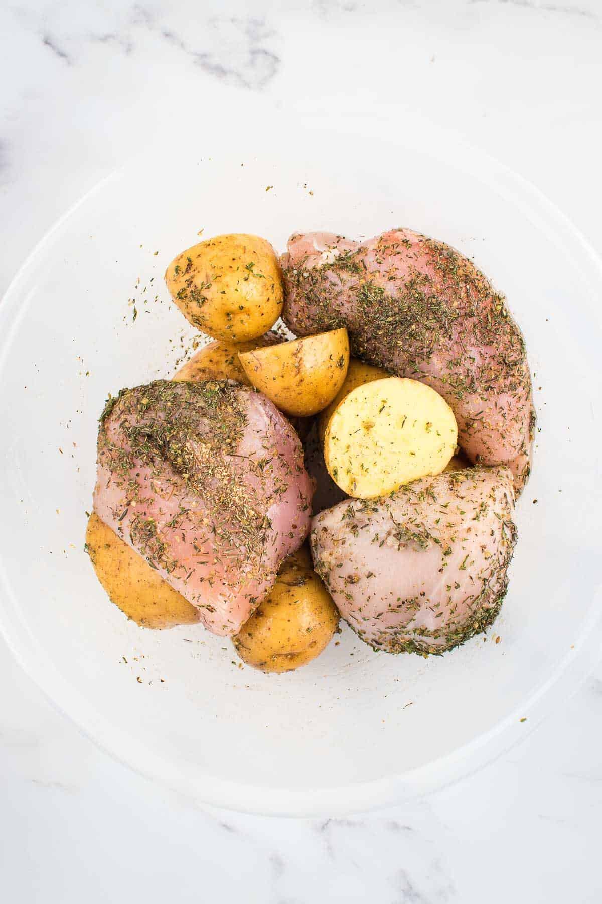 Raw chicken breasts, potatoes and seasoning in a bowl.