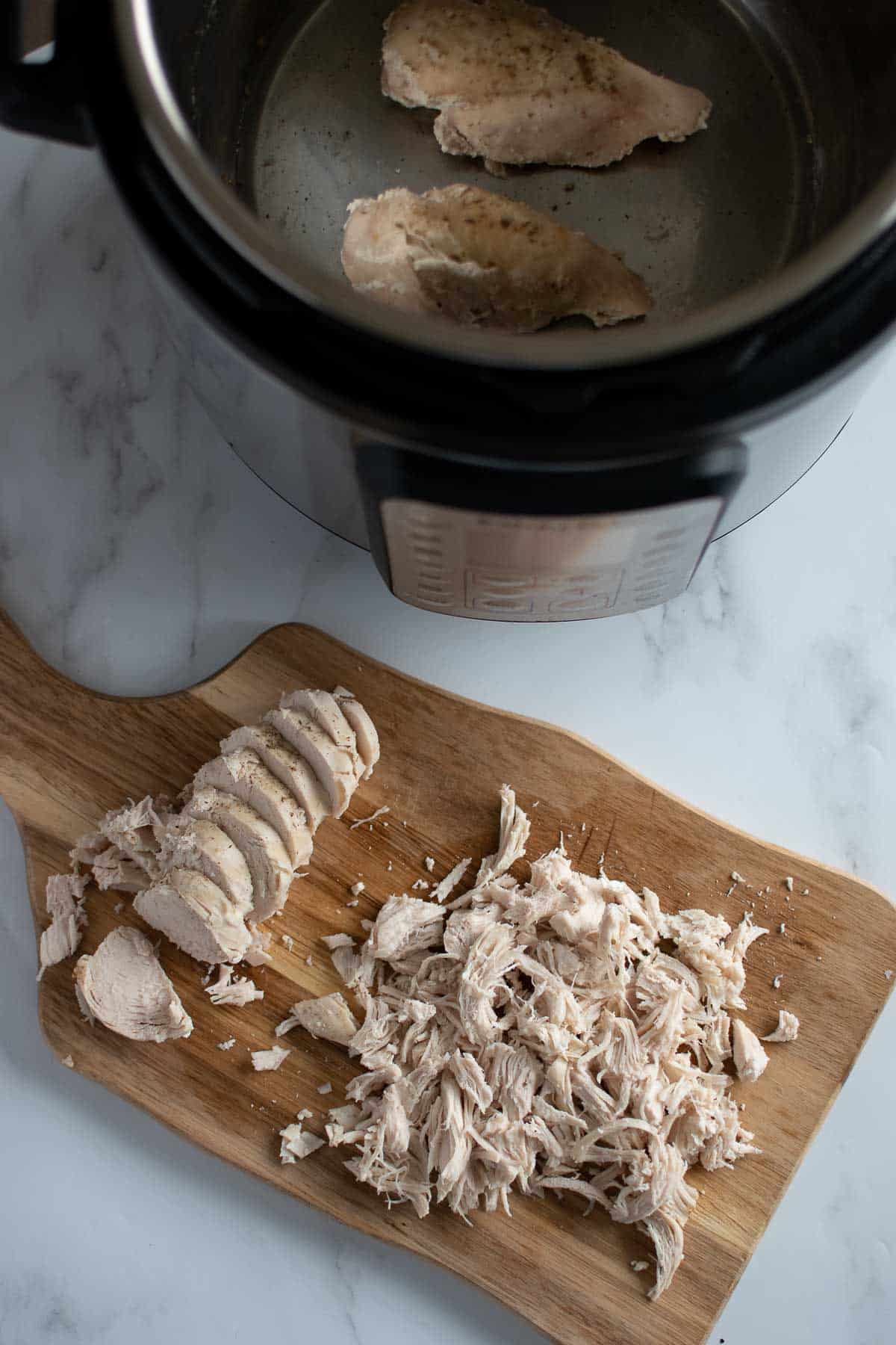 Sliced and shredded chicken breasts on a chopping board, with an instant pot with cooked chicken in the back.