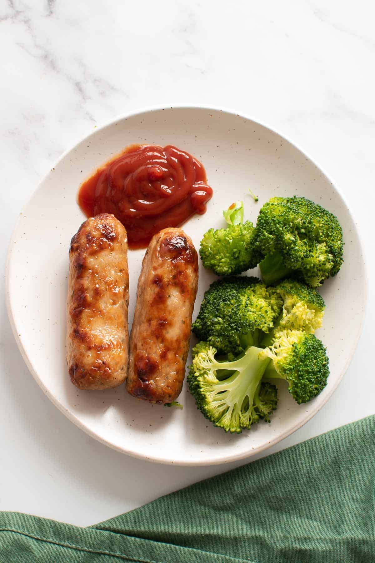 Cooked sausages on a plate with ketchup and broccoli.