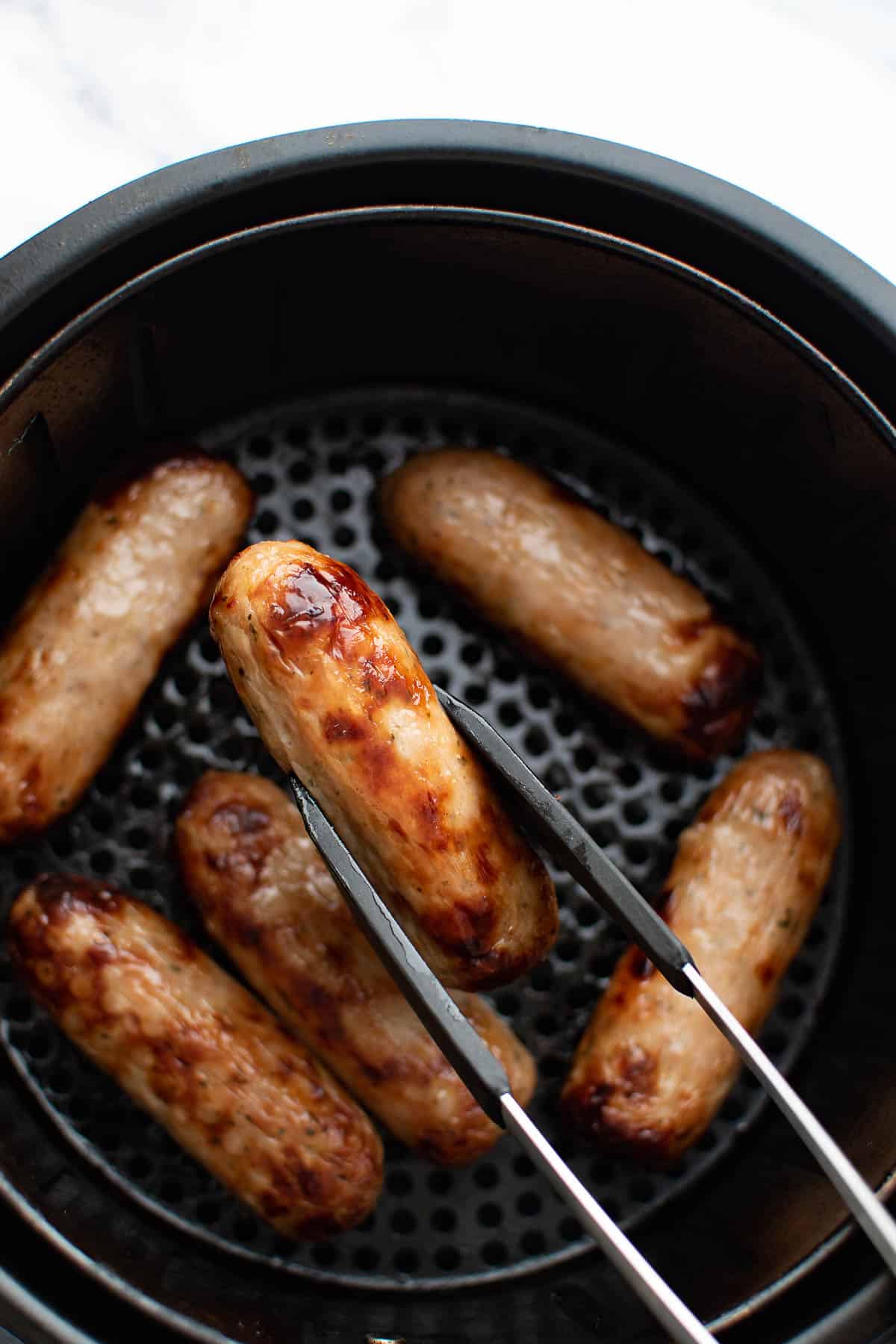 Sausages in an air fryer, with tongs lifting up one of the sausages.