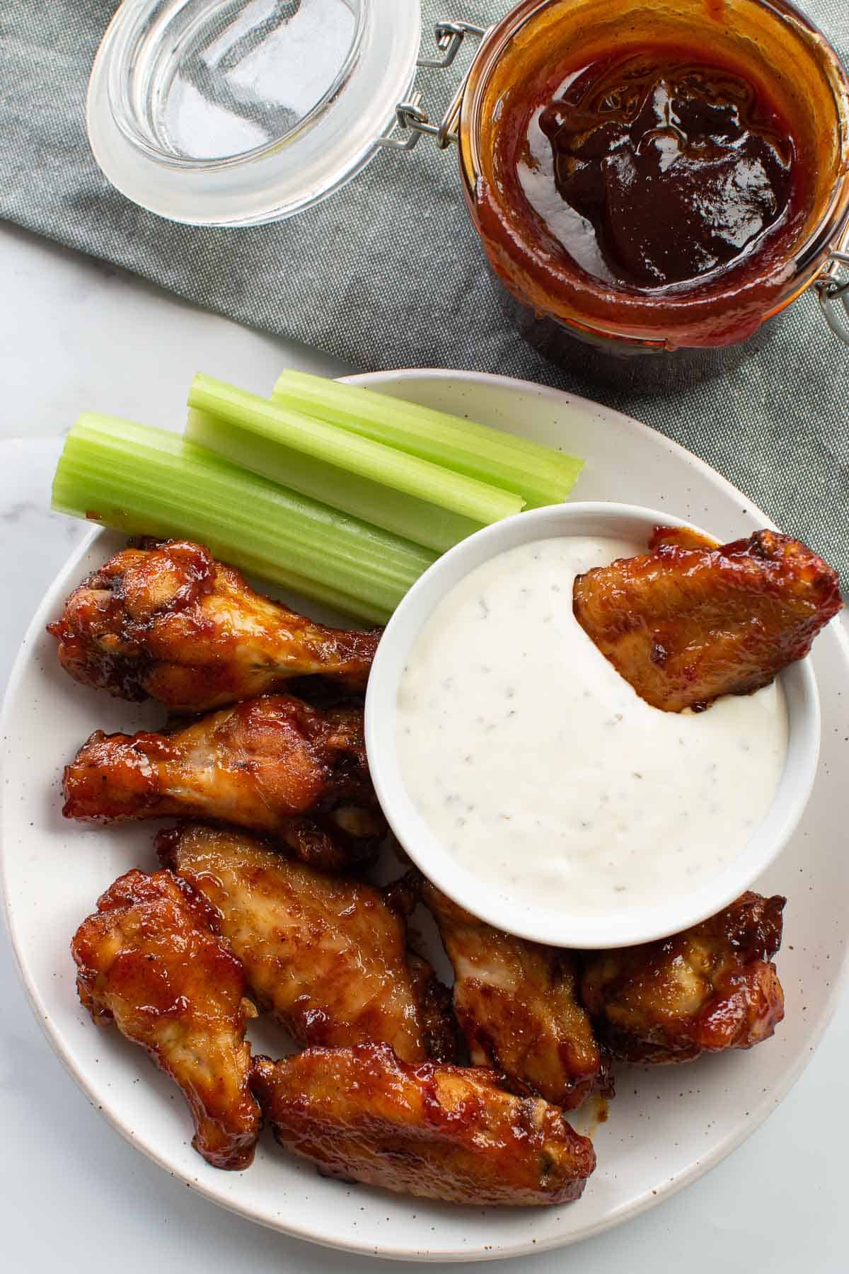 Baked BBQ chicken wings on a plate with a bowl of ranch and celery sticks.