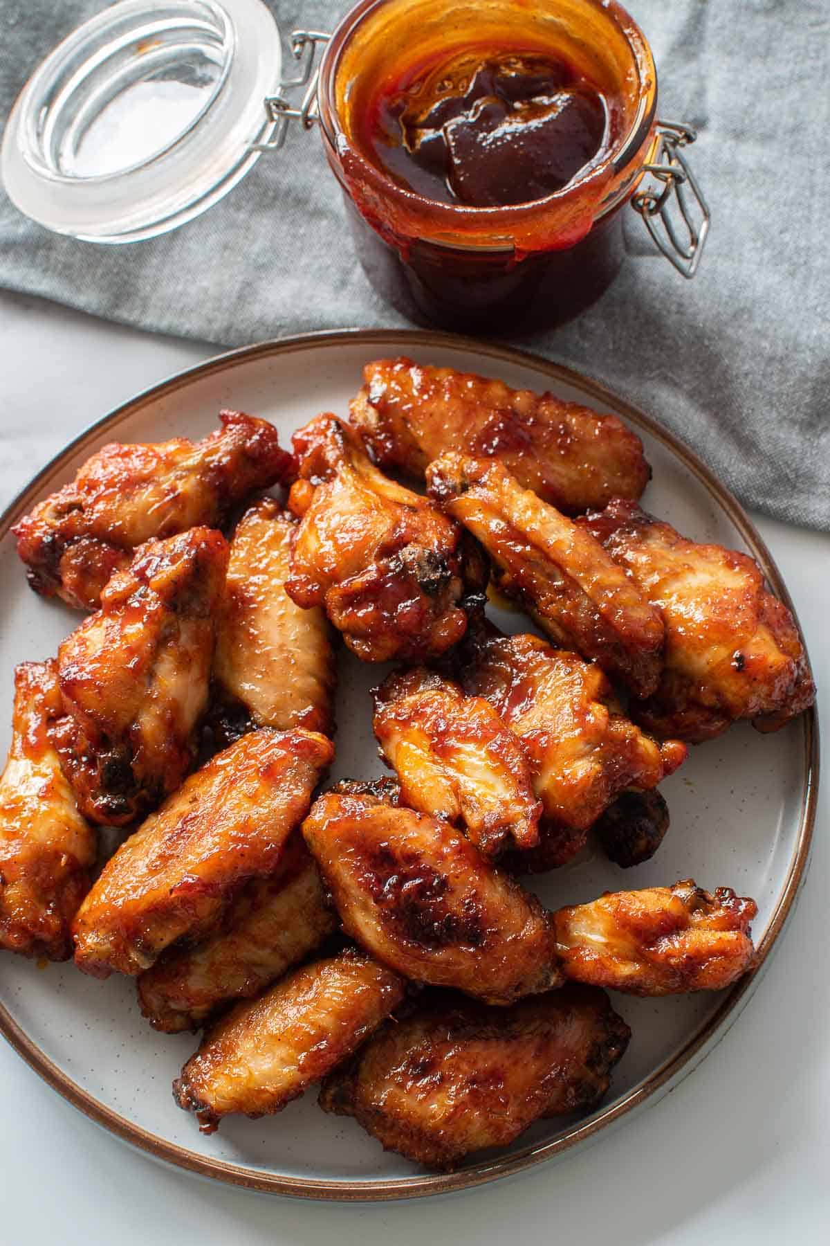 A plate full of baked BBQ wings, with a jar of BBQ sauce on the side.