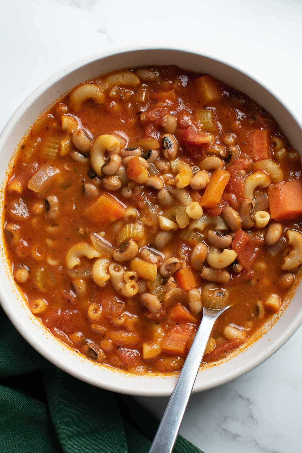 A bowl of tomato soup with pasta and black eyed peas, with a spoon submerged into it.