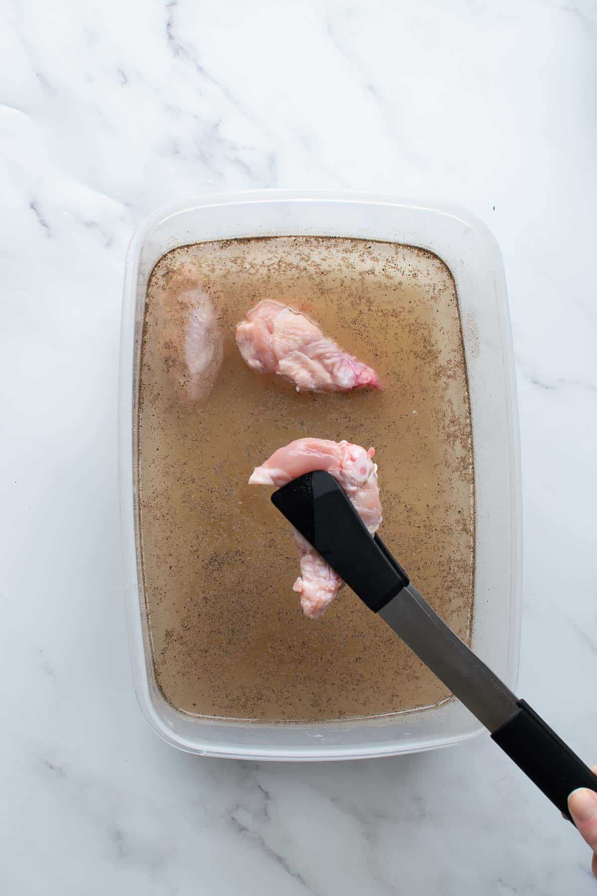 A container of brine, with tongs adding chicken wings.