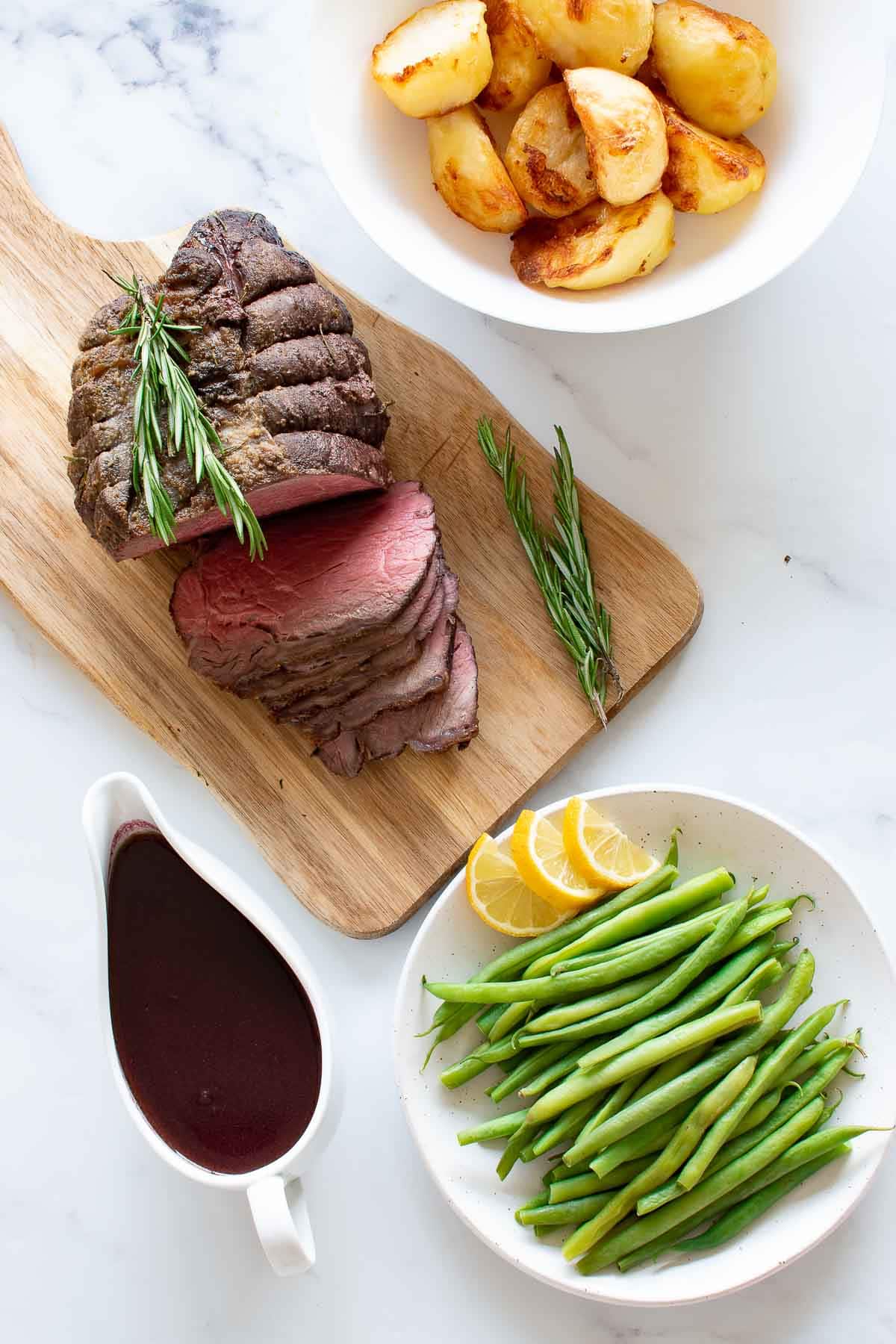 uma mesa com rosbife, feijão verde, batata assada e jus de vinho tinto.