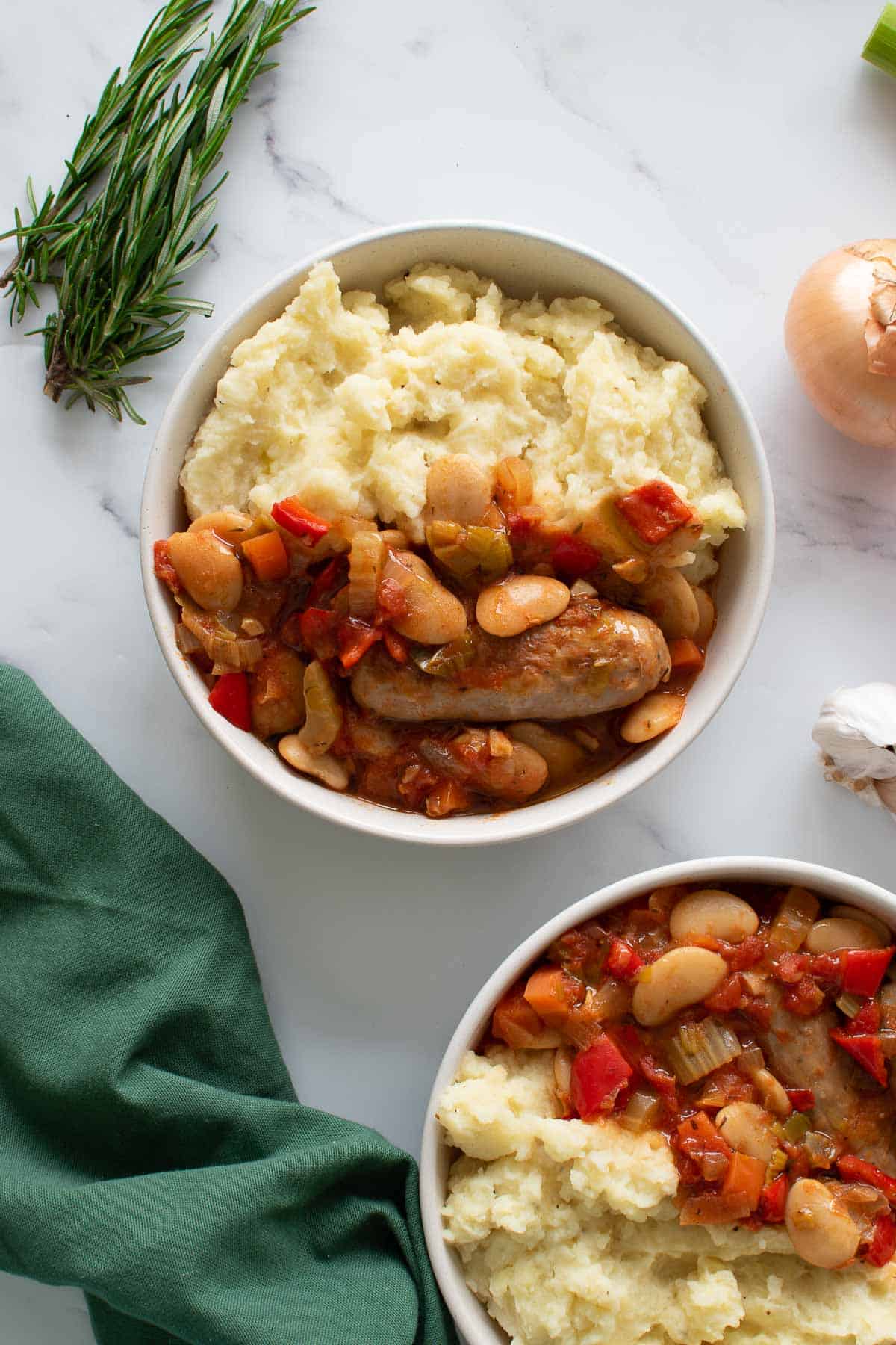 Two bowls of sausage stew, with ingredients scattered in the background.