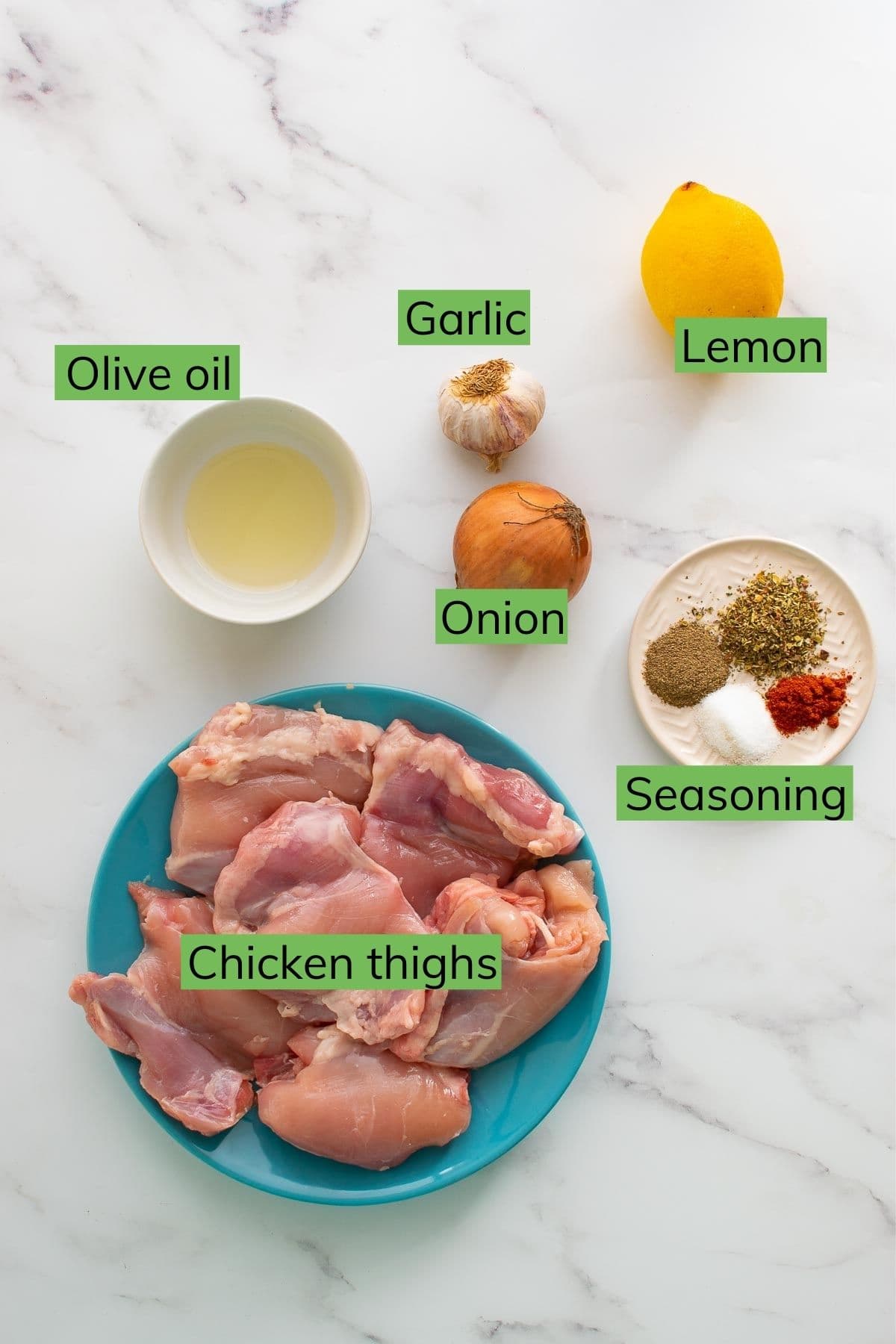 Chicken thighs, lemon, garlic, onion, seasoning and olive oil laid out on a table.