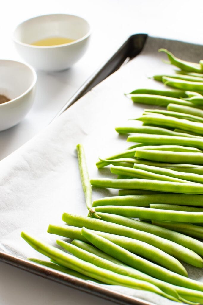 Green beans on a baking sheet, with seasoning and olive oil on the side.