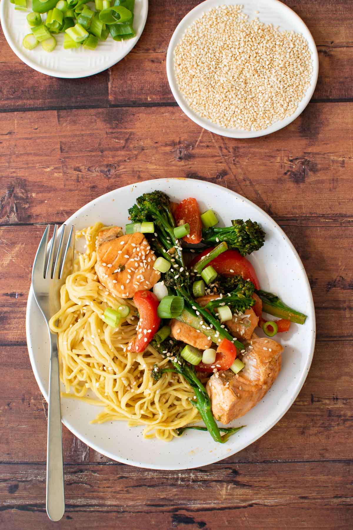 A plate with salmon and broccoli stir fry with noodles. Sesame seeds and scallions on the side.