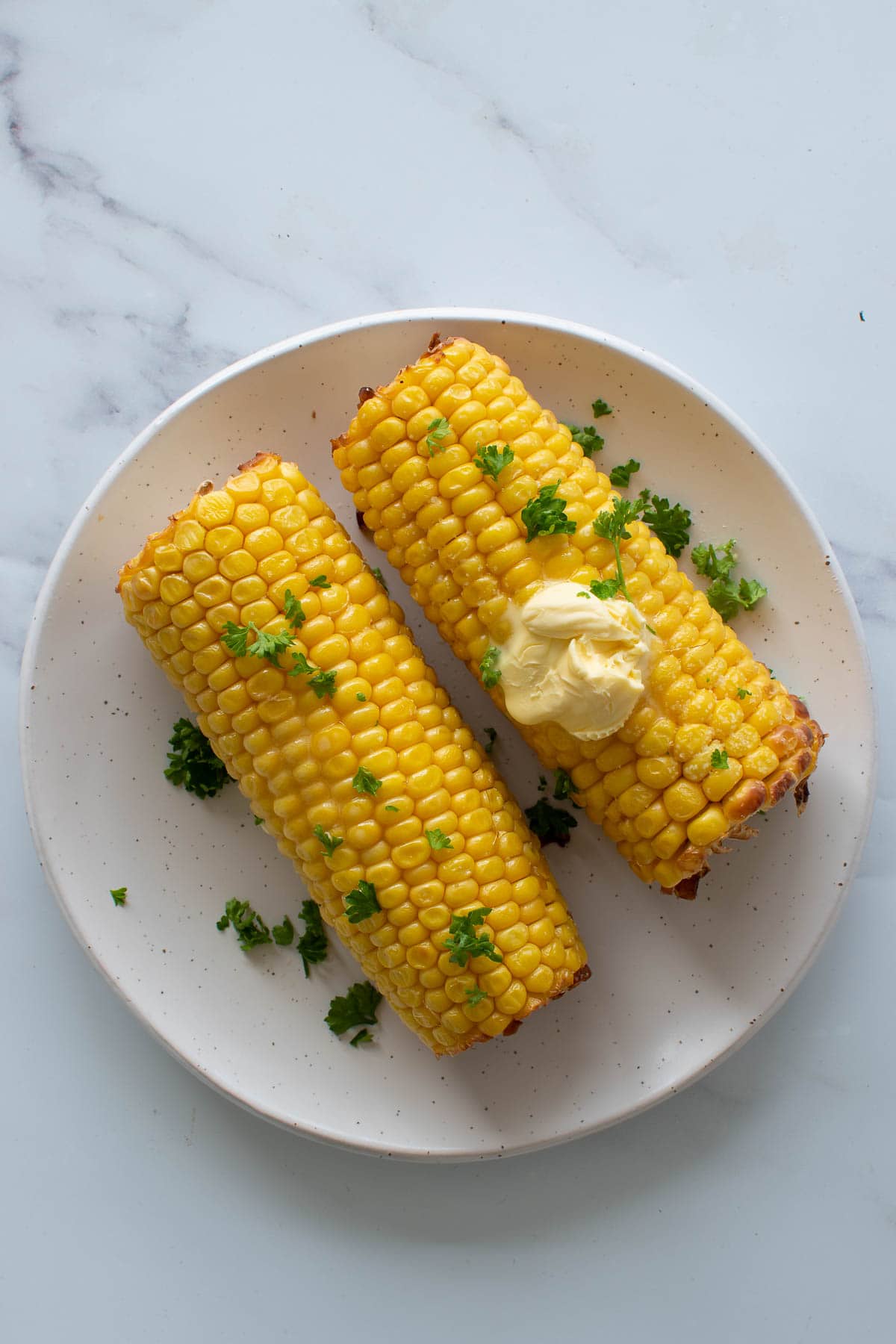 Two roasted cobs of corn on a white plate with parsley and butter on top.