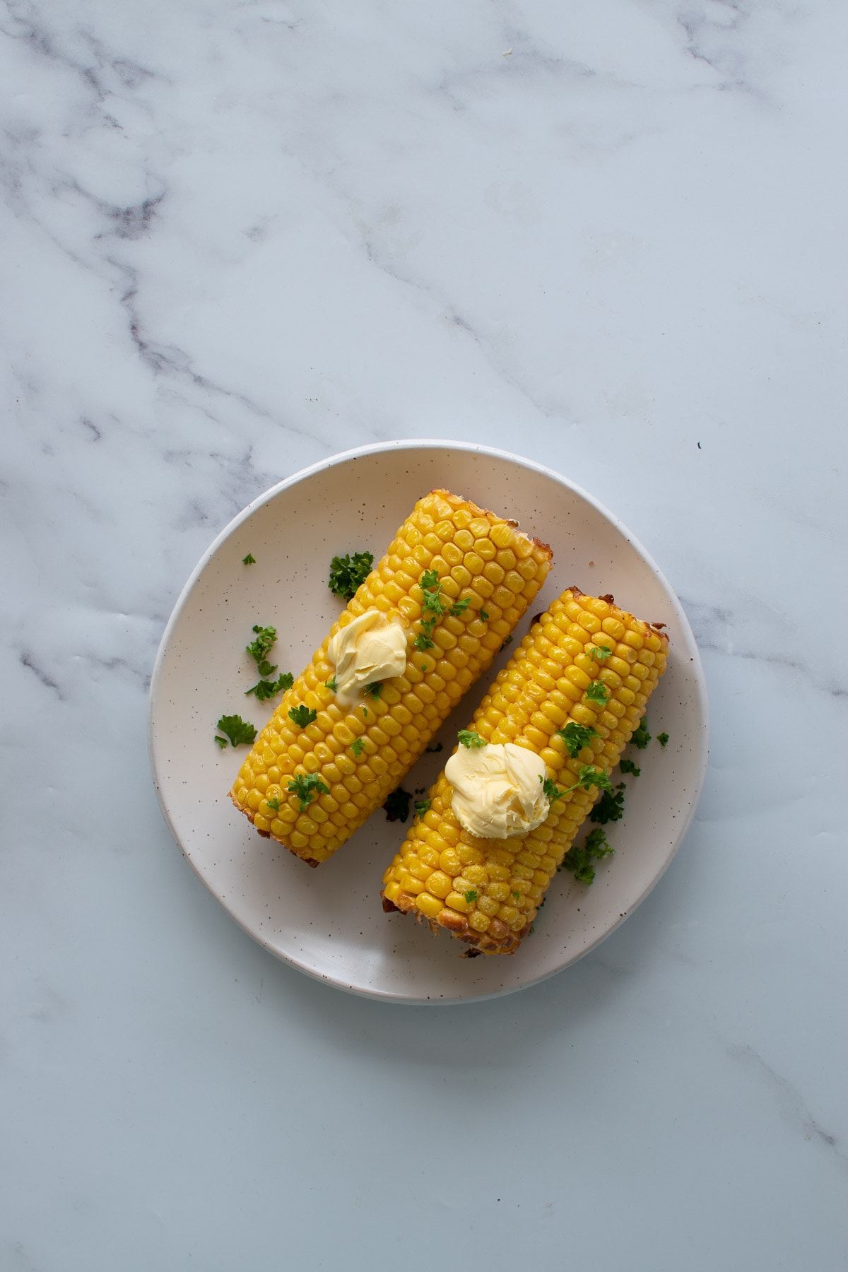 Two roasted cobs of corn on a white plate with parsley and butter on top.