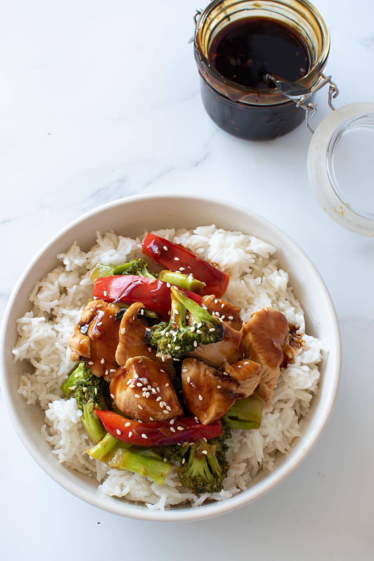 A bowl of rice and chicken stir fry, with a jar of teriyaki sauce in the background.