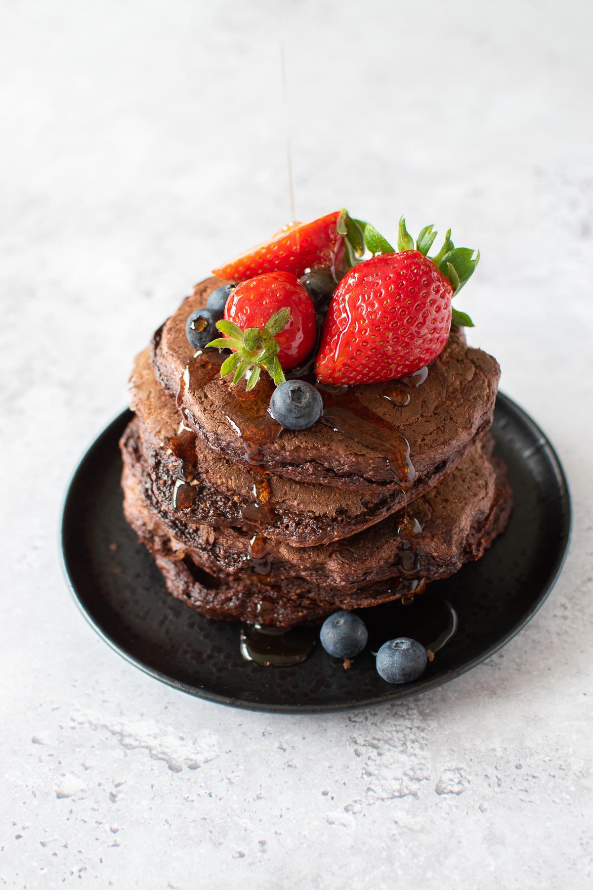 A stack of chocolate pancakes with strawberries on top, and maple syrup drizzling over it.