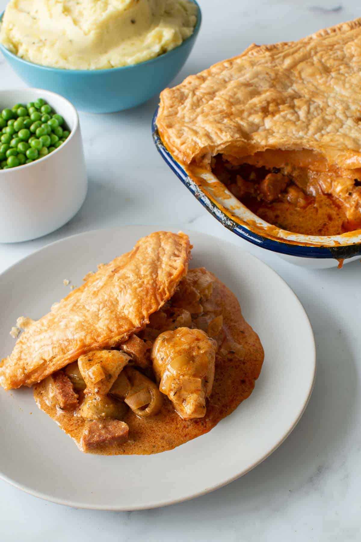 A plate of chicken and chorizo pie, with the pie, mashed potatoes and peas in the background.