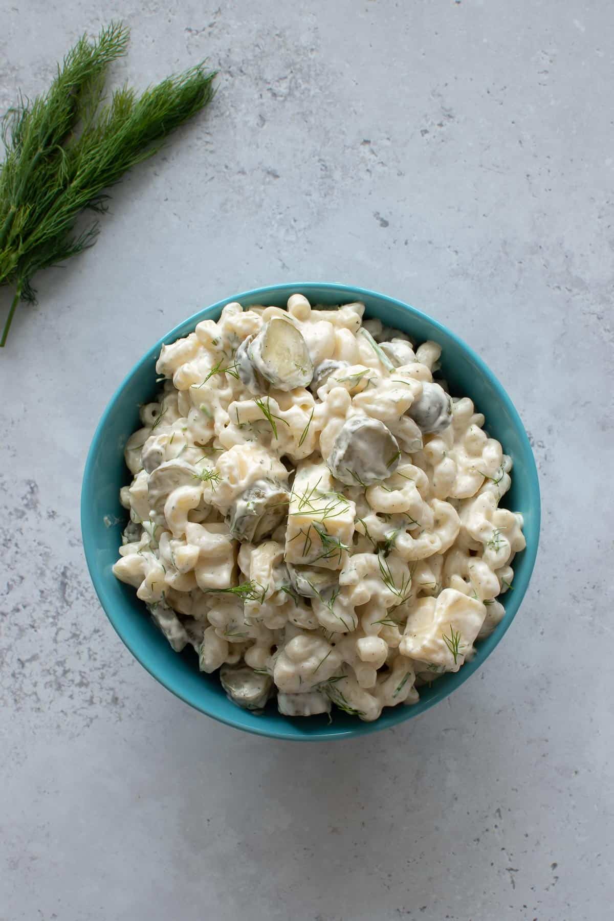 A bowl of pasta salad with cheddar and pickles, with dill on the side.