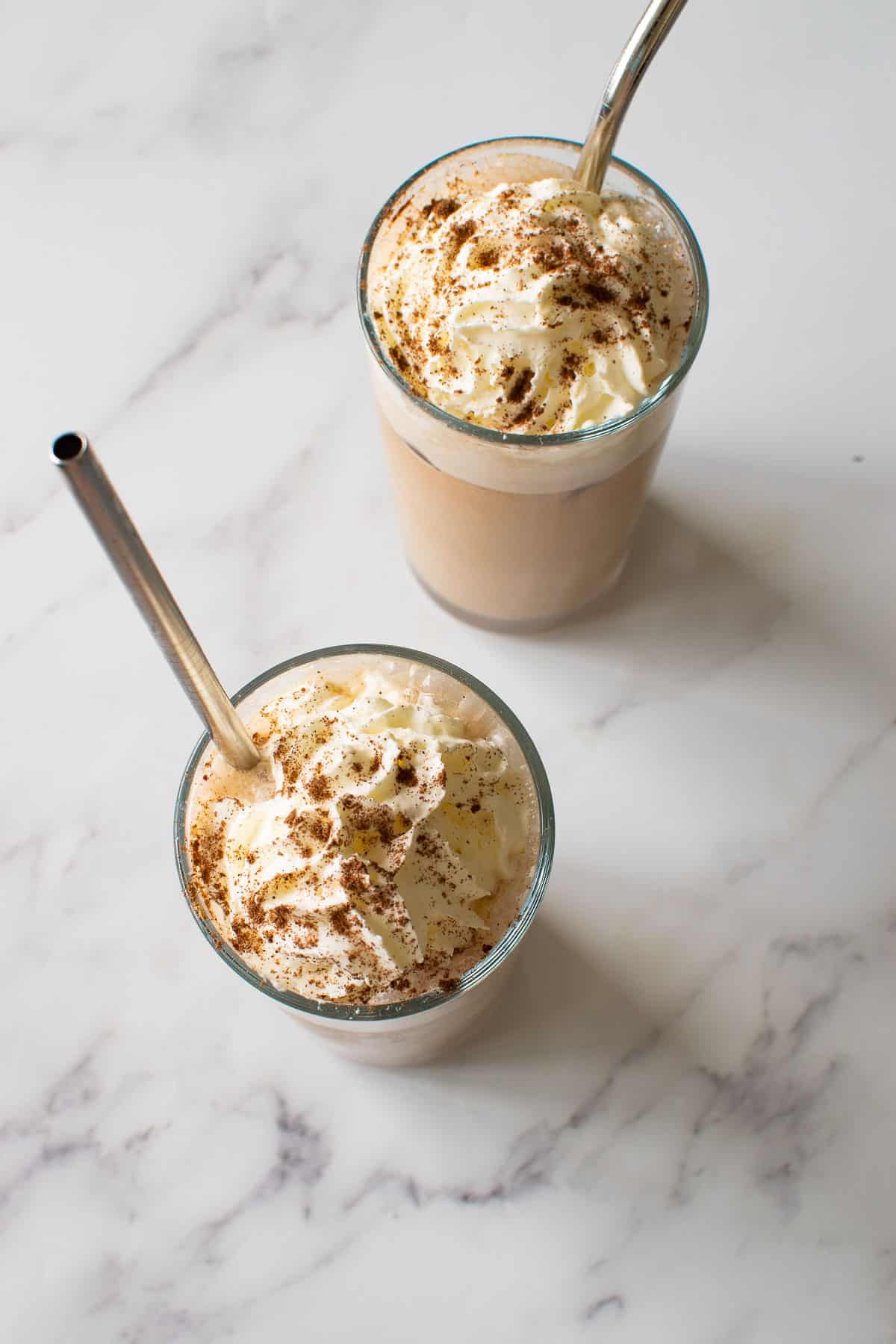 Two glasses of iced latte with whipped cream on a marble table, with metal straws inserted.