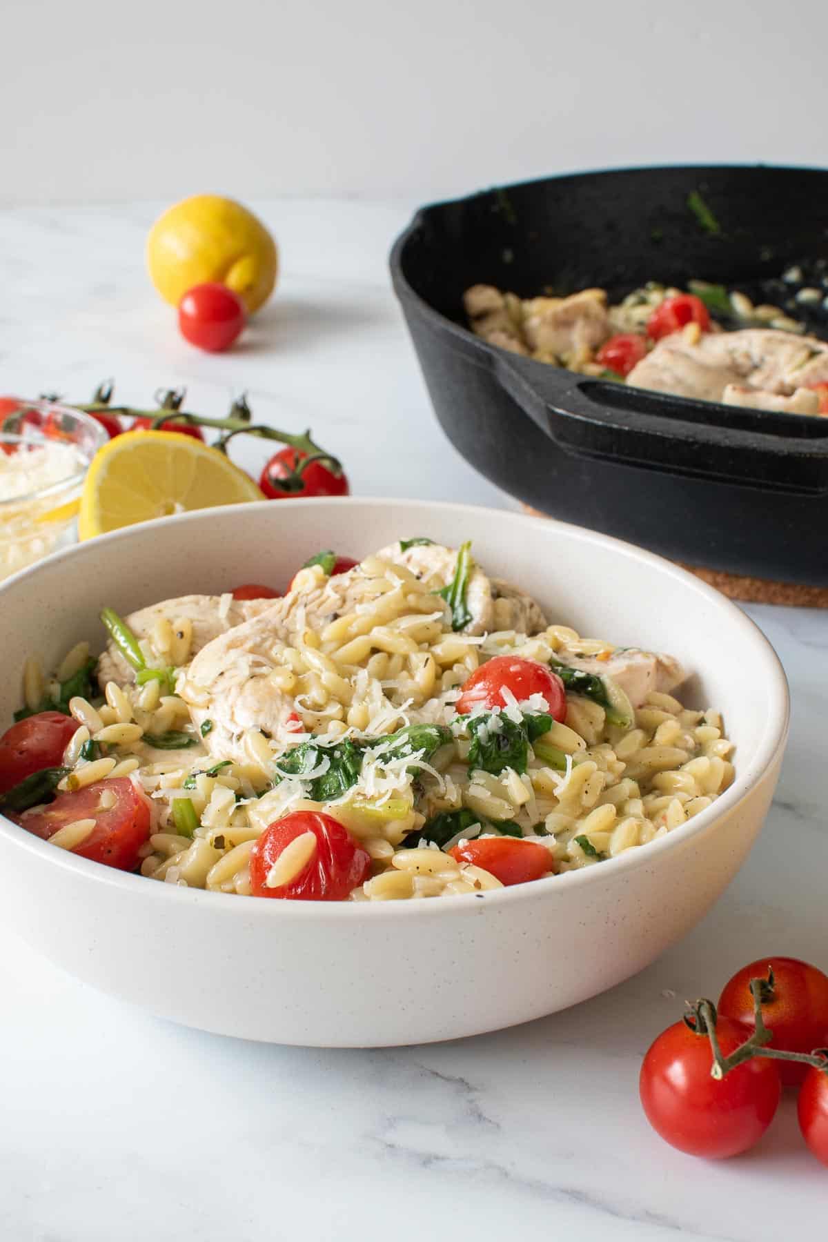 A bowl of lemon chicken with orzo and tomatoes, with a cast iron skillet in the background.