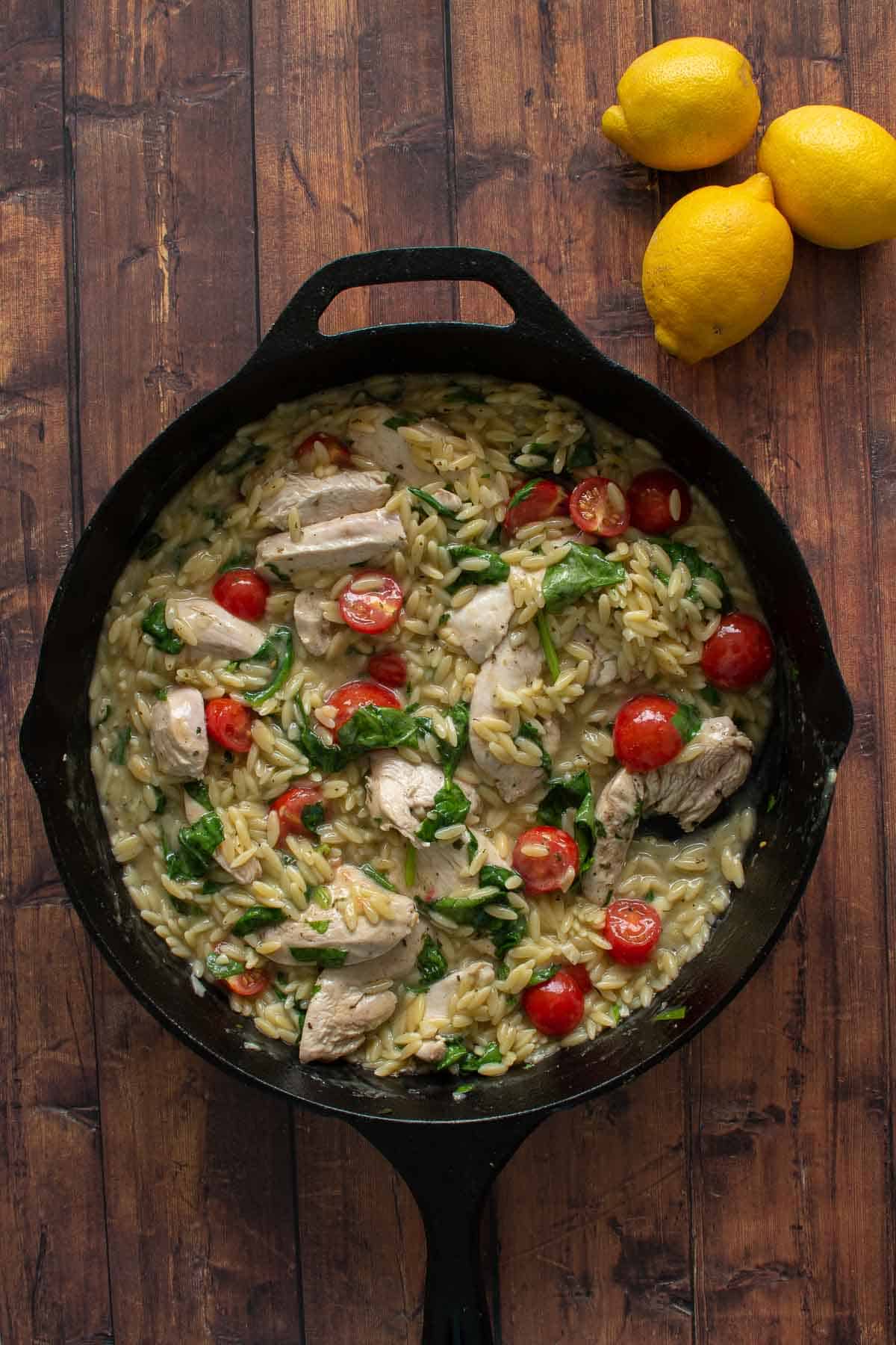 A cast iron skillet with lemon, orzo, chicken, spinach and tomatoes.