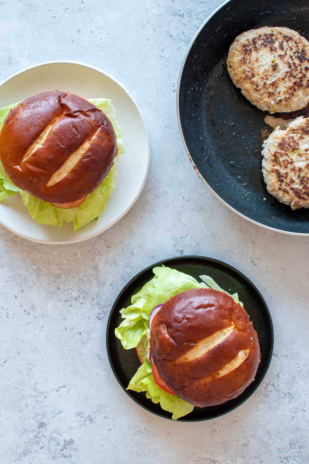 Two plated pork and apple burgers, with more burger patties in a frying pan on the side.