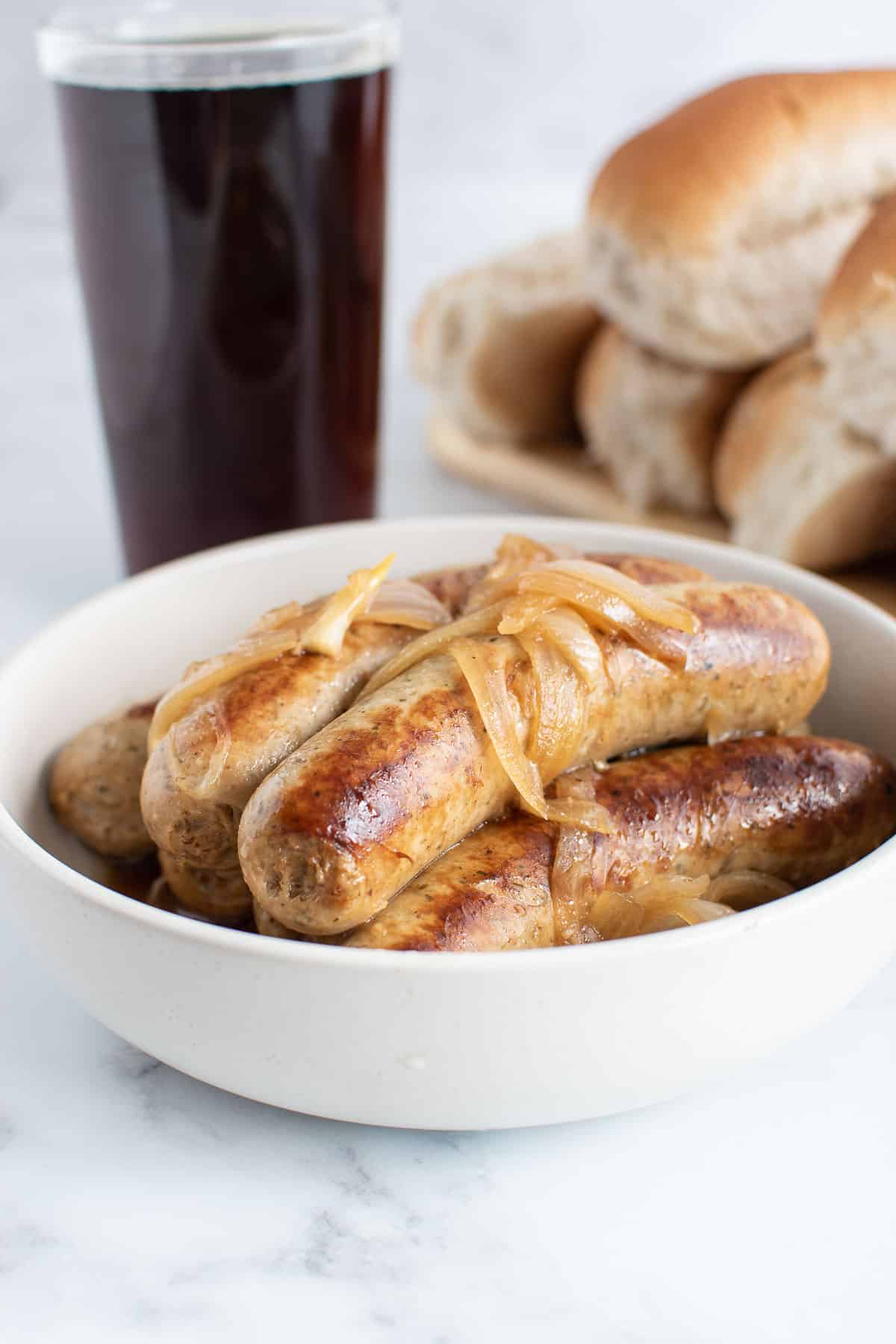 Bratwurst sausages in a bowl, with beer and hot dog buns in the background.