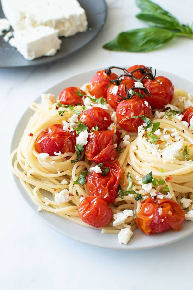 Roasted cherry tomatoes and spaghetti, with feta cheese and basil in the background.