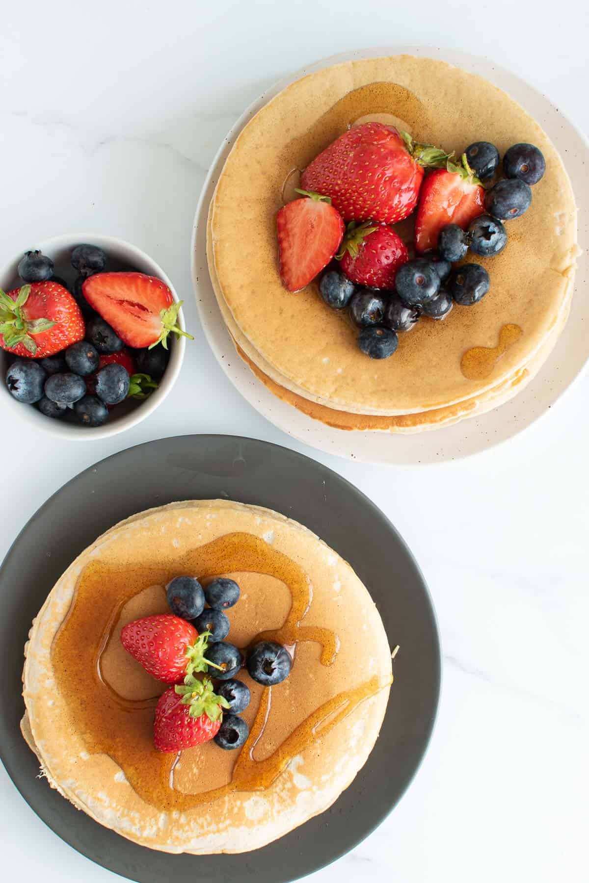 Two stacks of oat milk pancakes with fresh berries and syrup.