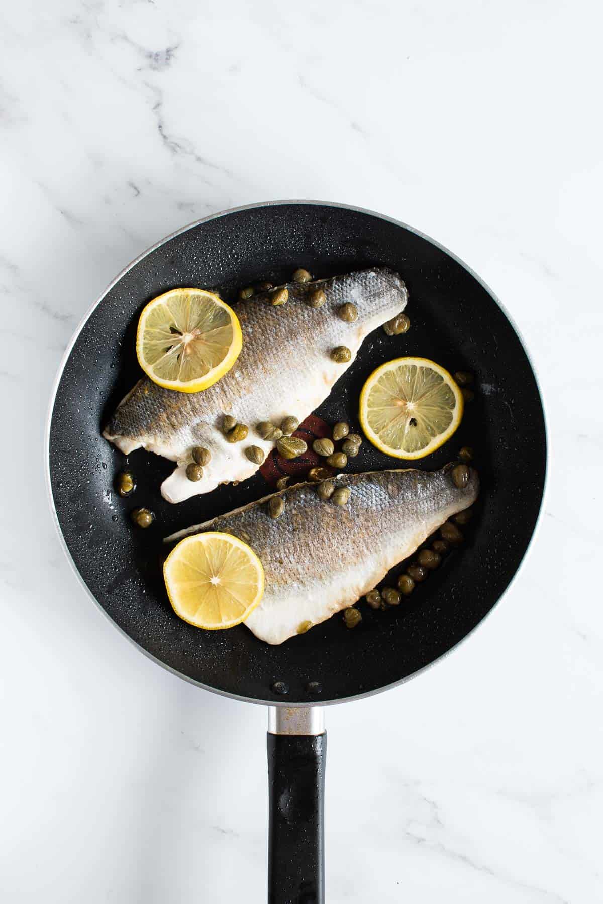 A frying pan with sea bass fillets, lemon and capers.